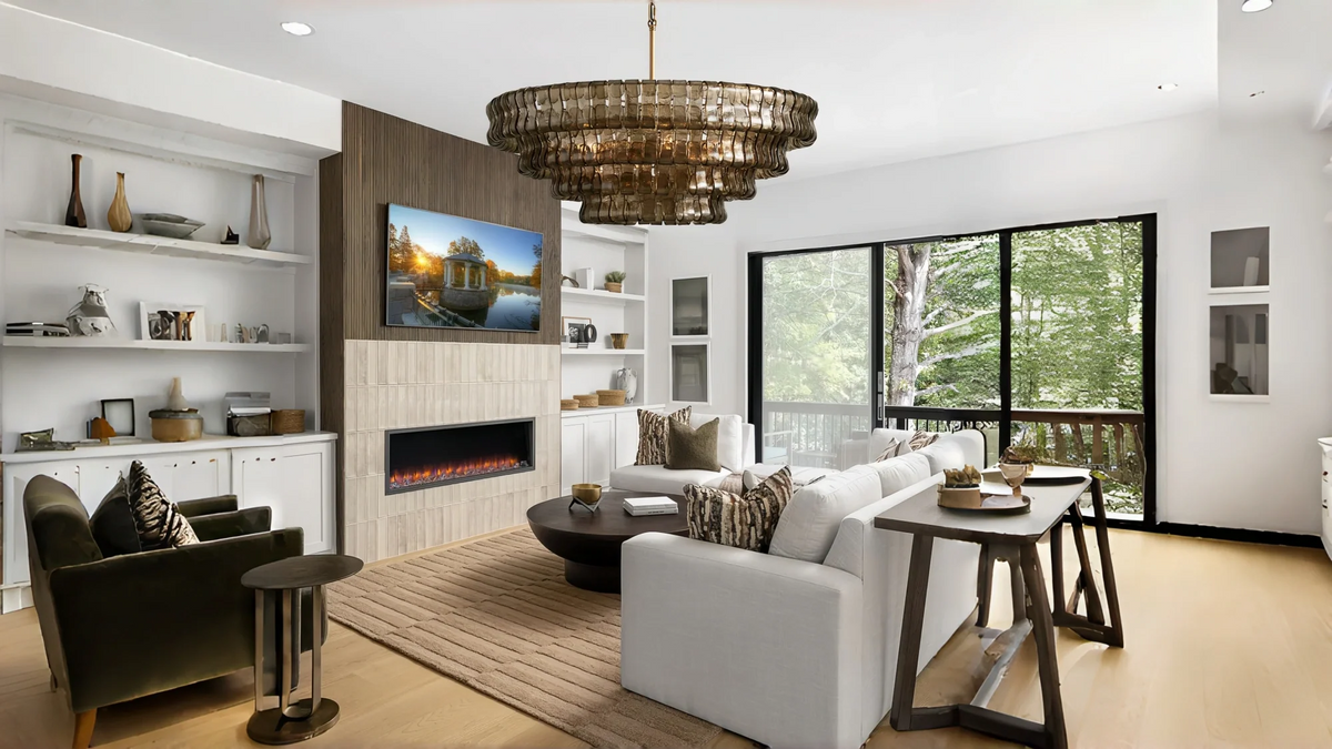 A modern living room interior with a large round chandelier hanging above a fireplace, white sofas, a wood dining table, built-in shelving, and floor-to-ceiling windows overlooking trees.